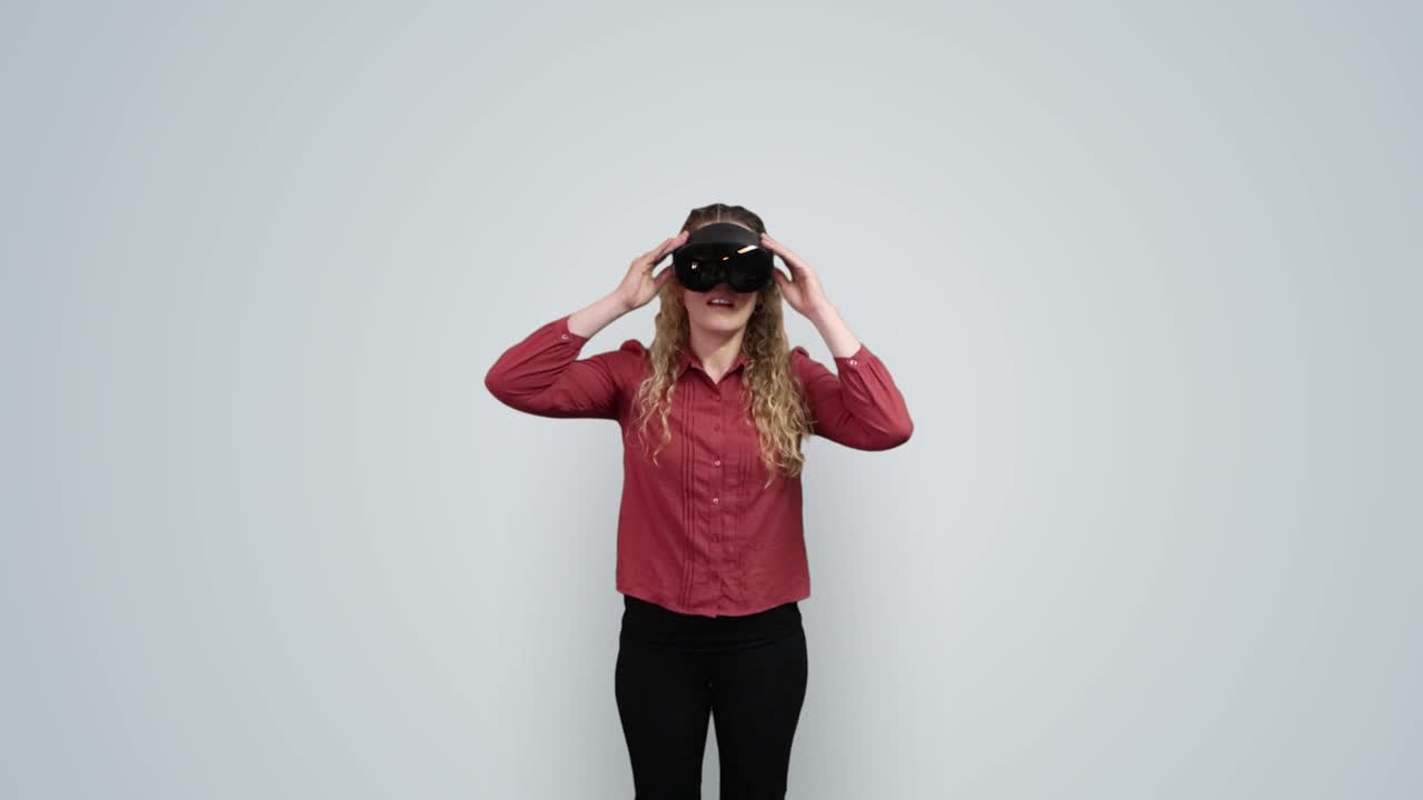 A woman in a red blouse lifts a black VR headset onto her face with her hands, against a plain grey backdrop