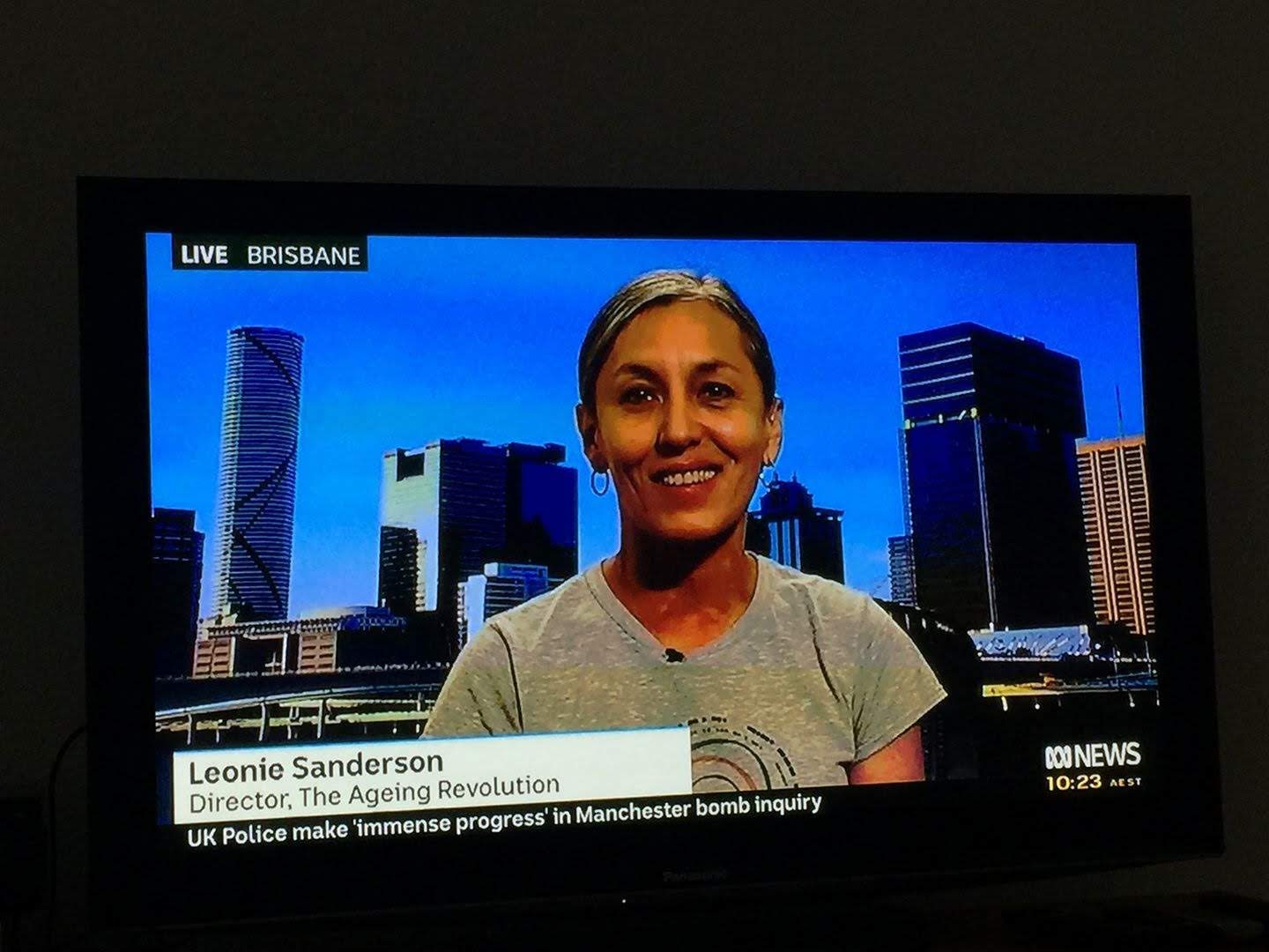 A television screen showing Leonie Sanderson in a grey t-shirt live on ABC News, the Brisbane skyline behind her and a caption naming her Director of The Ageing Revolution.