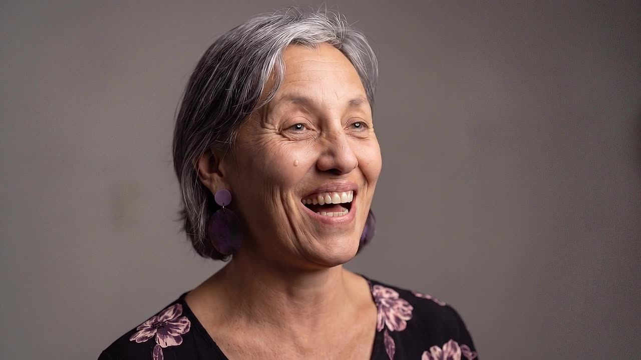PTR co-founder Leonie Sanderson laughing in a close studio portrait, purple disc earrings and a dark floral top against a plain grey backdrop.