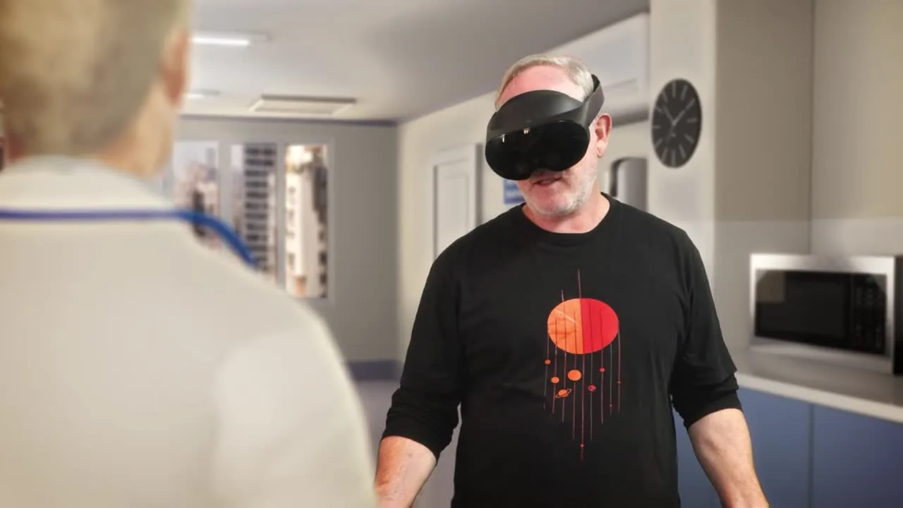 A man wearing a headset stands in a hospital staff room, a colleague with a lanyard in the foreground, rehearsing a moment on shift.