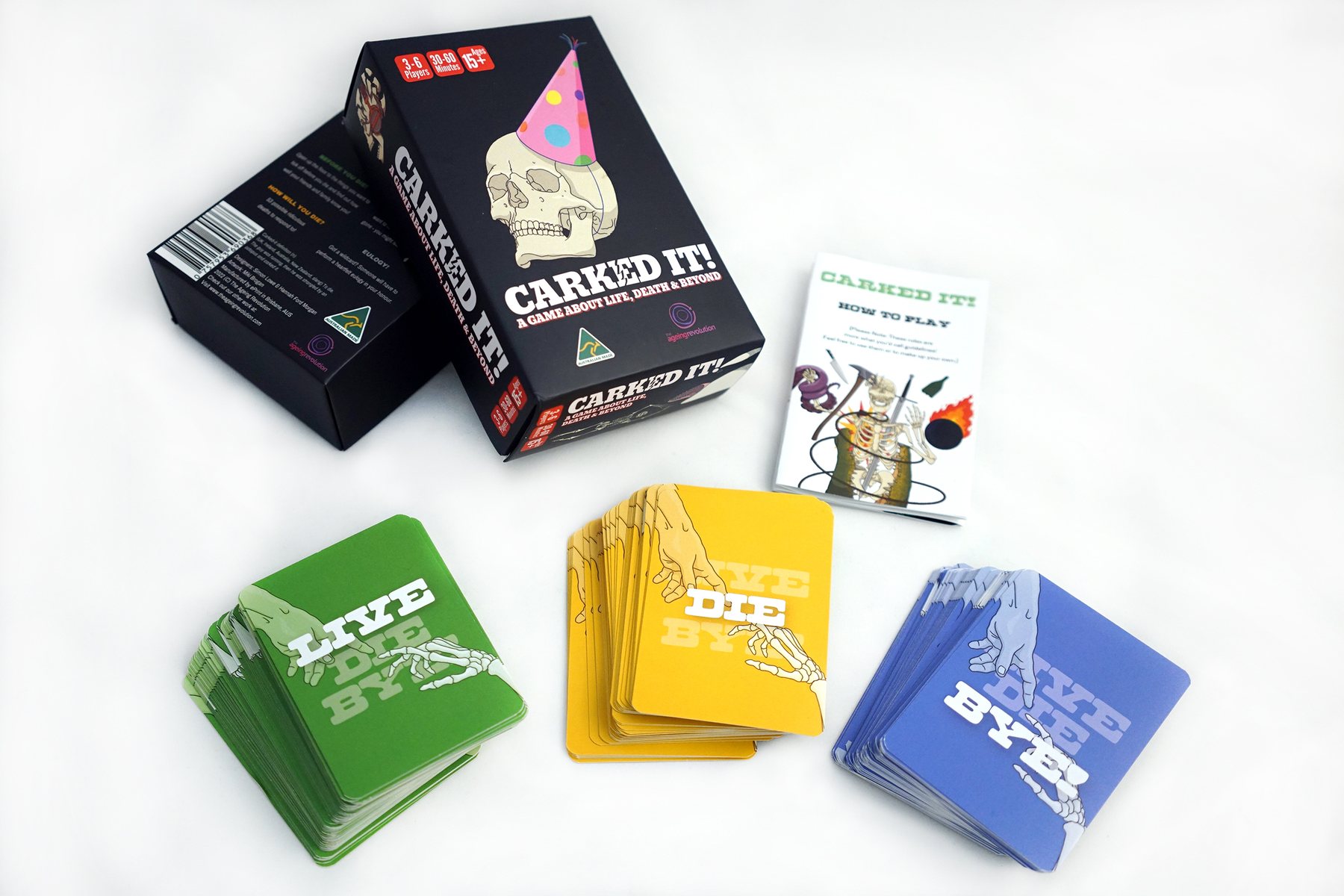 The Carked It! card game laid out on white: a black box with a skull in a party hat, a how to play booklet, and three stacked decks of green, yellow and blue cards.