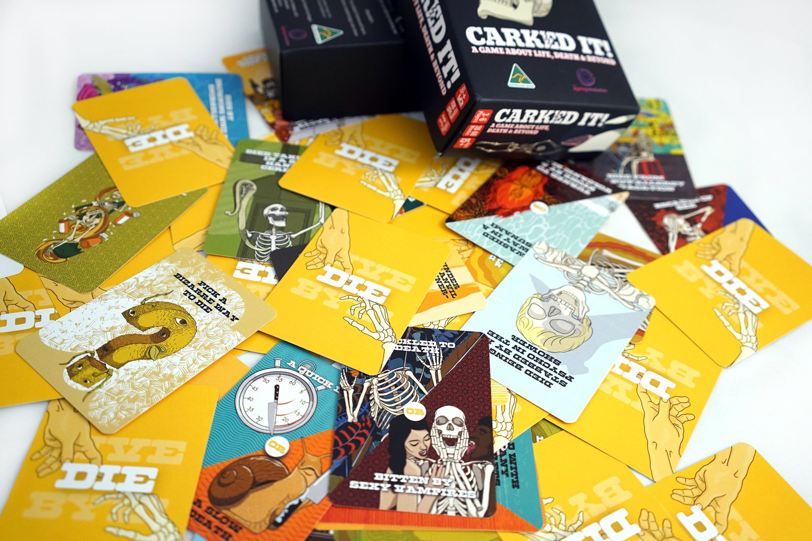 Yellow cards from the DIE deck fanned across a table beside the box, each illustrated with a different way of dying.
