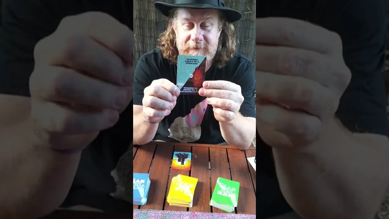 A man in a wide-brimmed hat holds a Carked It! card up to camera above a wooden table with the decks spread out below him.