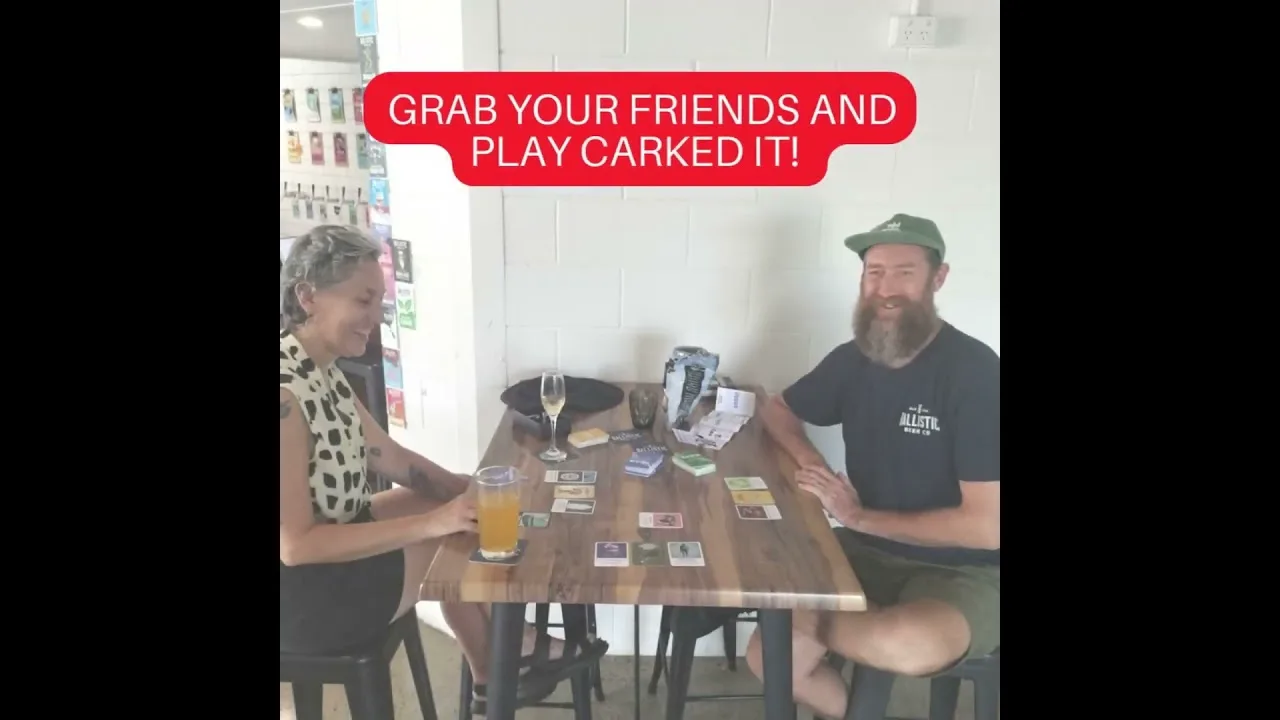 Two people sit at a wooden pub table with beers and the Carked It! cards spread between them, looking up at the camera.