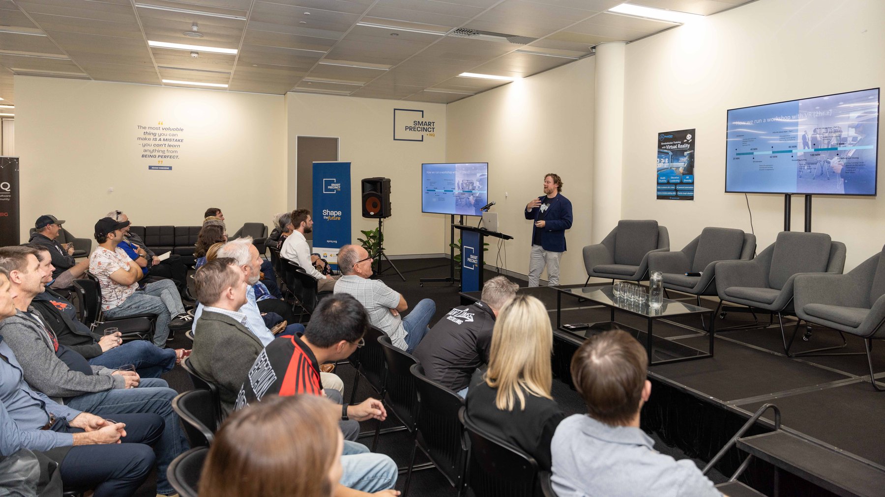 A seated audience watches a PTR presenter explain a slide titled 'How we run a workshop with VR' at a community event sharing how immersive training sessions are run.