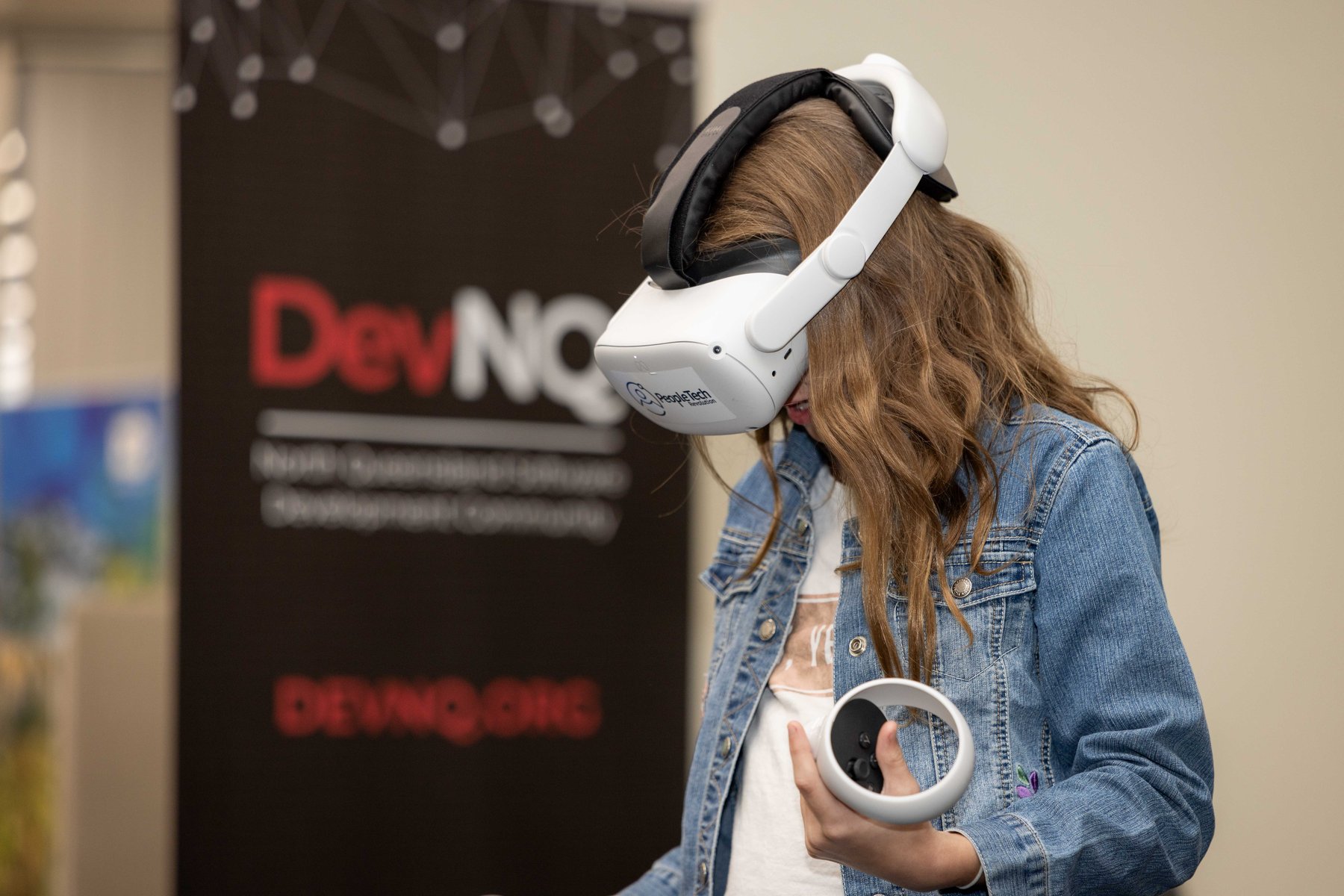 A young woman in a denim jacket wears a PeopleTech Revolution branded VR headset, holding a controller, trying an immersive training demo at a PTR community event.