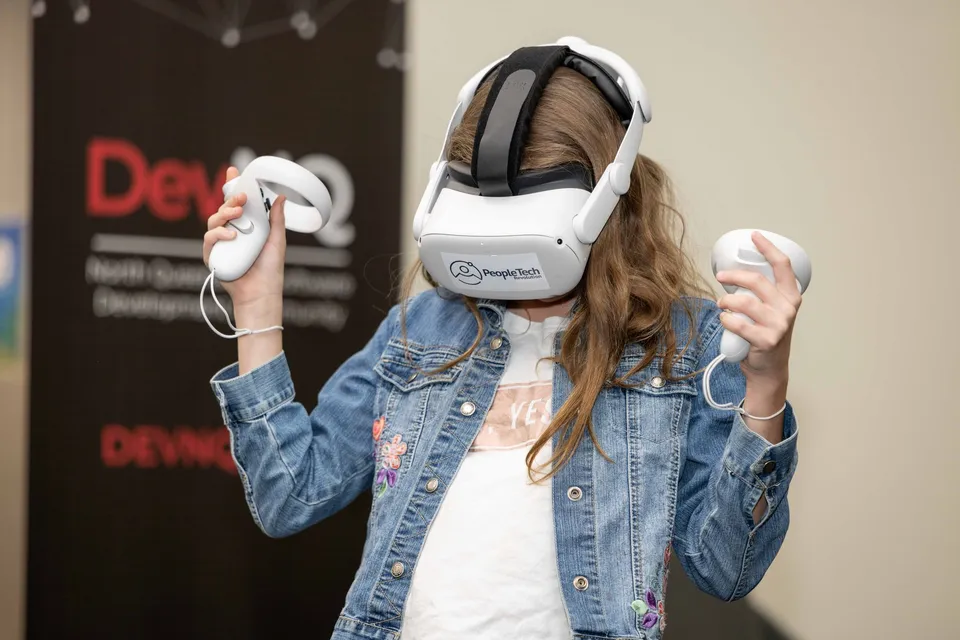 A young woman in an embroidered denim jacket wearing a PeopleTech Revolution branded headset raises both controllers, trying immersive tech hands-on at a PTR community event