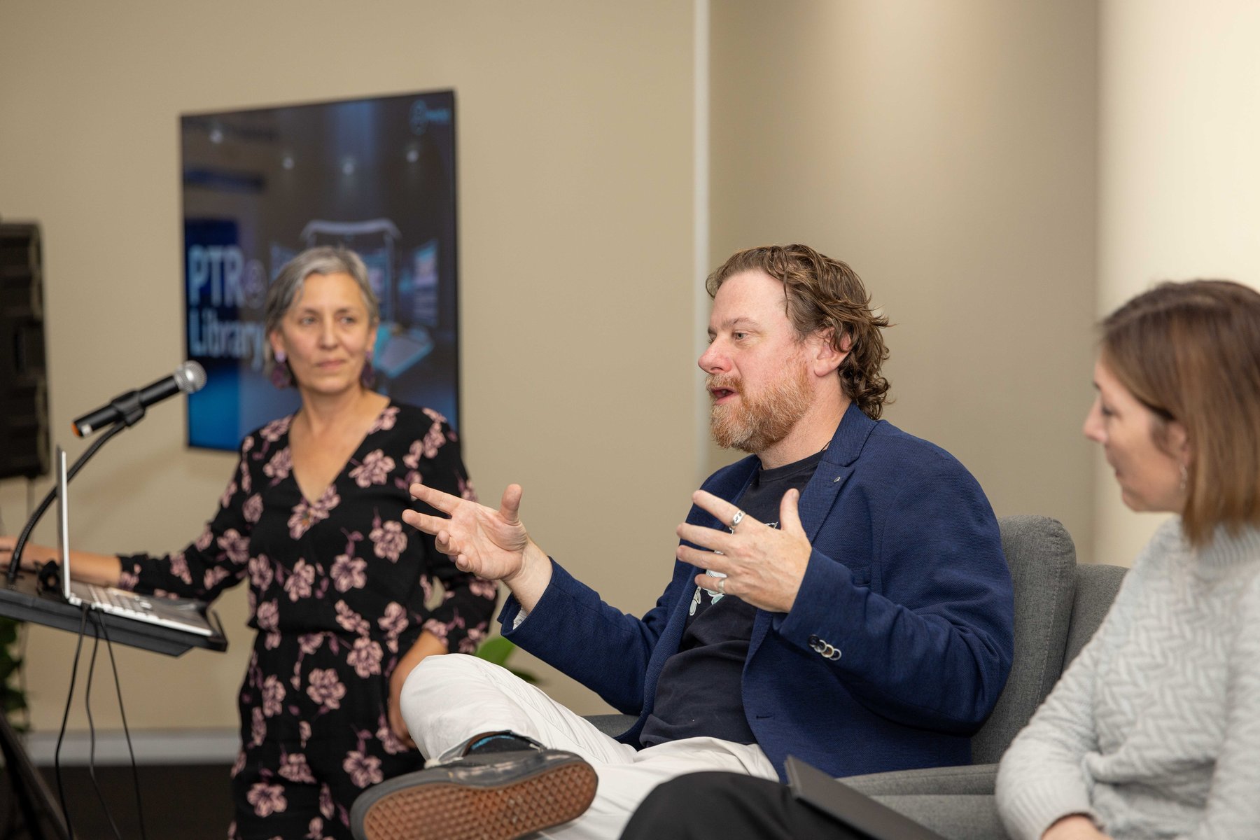 Leonie Sanderson stands at a lectern with a laptop as a bearded panellist gestures while speaking, another panellist listening beside him, at a PTR community event