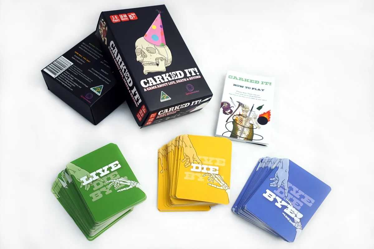 The Carked It! card game laid out for its Australian Good Design Award entry: the black box with a party-hat skull beside the LIVE, DIE and BYE decks.