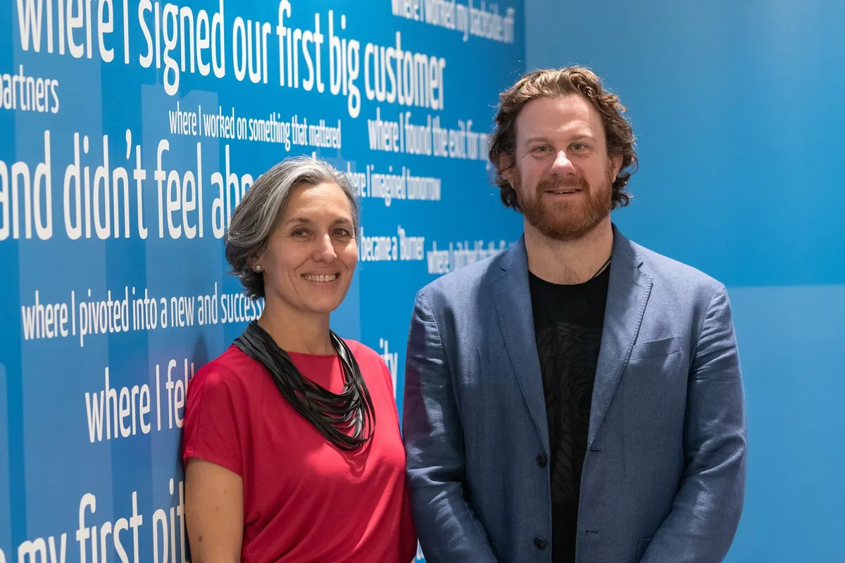 Leonie Sanderson in a red top and Simon Lowe in a blue jacket stand side by side in front of a blue wall printed with white phrases that each begin 'where I'.