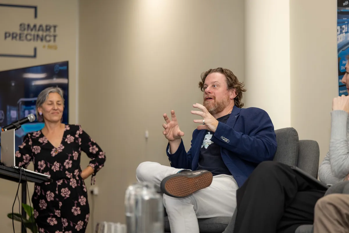 Leonie Sanderson speaking into a microphone beside Simon Lowe, who is seated and gesturing, in front of a Smart Precinct NQ banner.