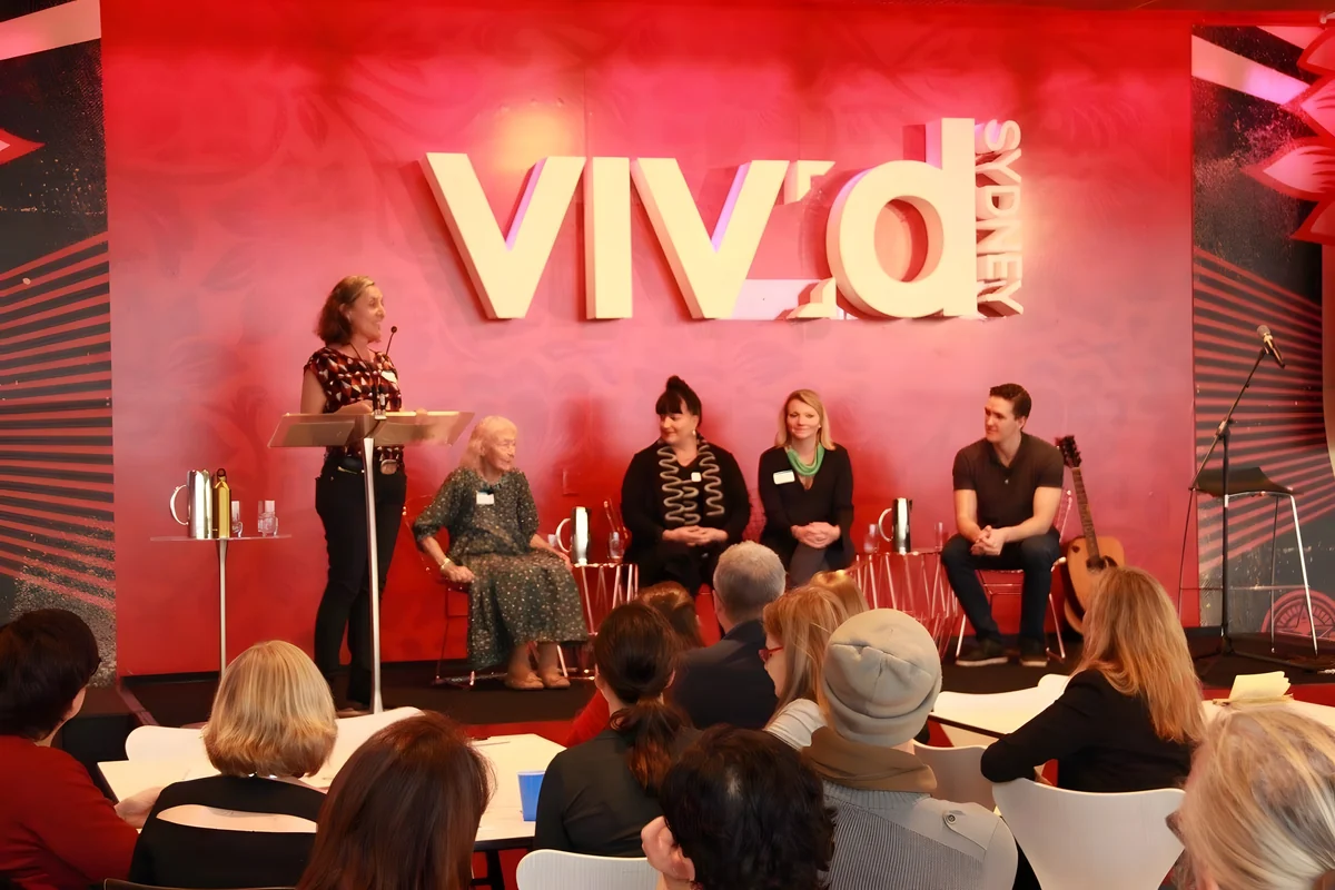 A red-lit Vivid Sydney stage: a speaker at a lectern on the left and four panellists seated in front of the Vivid sign, with an audience in the foreground.
